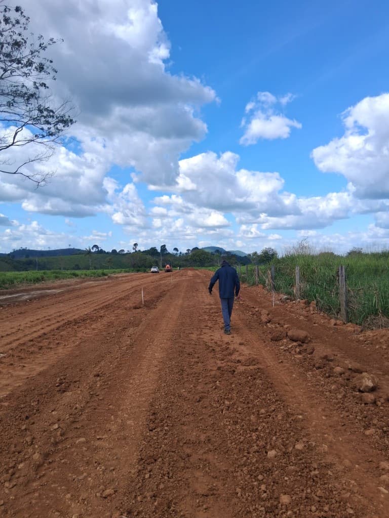 Via de terra em construção com caminhonete e rolo compactador ao fundo